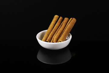 Cinnamon sticks in a white plate on a dark table with reflection. Close-up