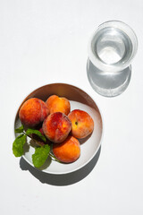 From the second, ripe peaches in a white town stand on a table in daylight next to a glass of water.