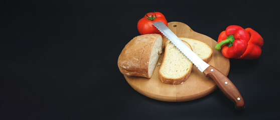 Knife slicing freshly baked loaf of bread on wood boaed with tomato and pepper on black background.
