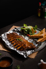 Close up grilled bbq ribs on aluminium foil in grey circle ceramic plate with toast on black table