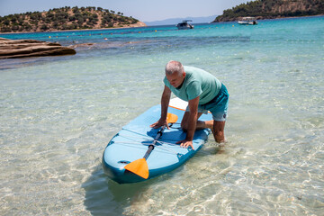 Sup surfing. Beautiful view of the sea with a mature man kneeling on a board with paddle in the water. Standup paddleboarding in summer