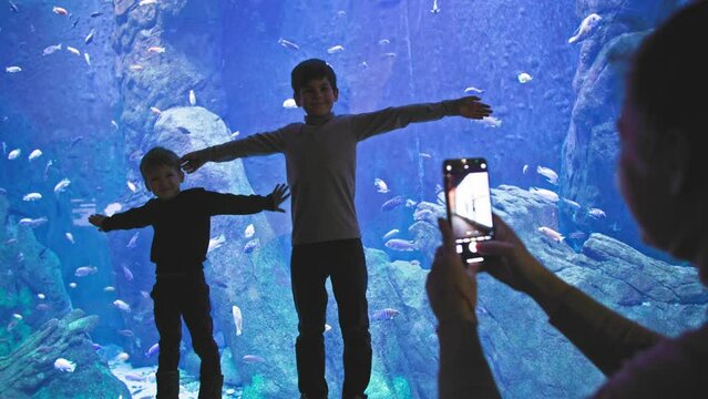 underwater world, woman with smartphone in hands is filming male children in front of a large aquarium with different fish in oceanarium