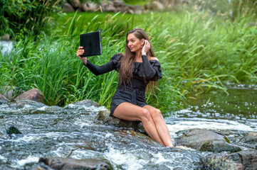 female sitting on a stone in the river and making a video call on a digital tablet