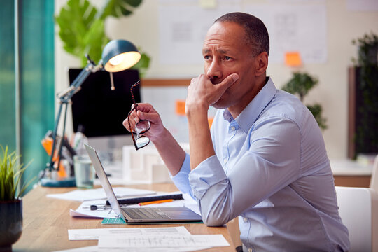 Stressed Mature Male Architect Working In Office At Desk On Plans For New Building