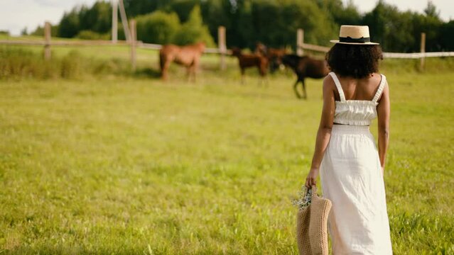 African-american woman walking in the field. Beautiful black female in white clothes and hat in horse range. Vintage smiling woman farmer lifestyle. African person in beautiful nature. 