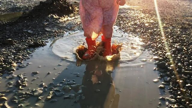 Little girl in rubber boots jumping in a puddle on a warm, sunny summer day