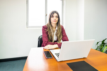 Young blonde business woman sitting at the head of the table in the office meeting room, with her computer and cell phone, looking at the camera