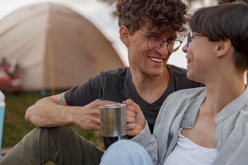 Portrait of young cheerful couple sitting at tent outdoor, laughing and having coffee in cups.