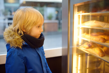 Fototapeta na wymiar A pretty child looks at a display case with freshly baked croissants in a European bakery on a cold and dark winter morning.