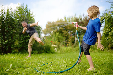 Funny little boy with his father playing with garden hose in sunny backyard. Preschooler child...