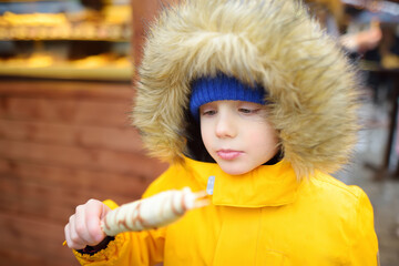 Little boy eating fruit in chocolate glaze on street Christmas market. Famous fruits, candy,...