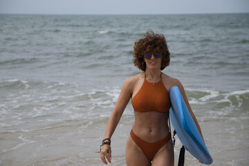 Attractive mature woman with curly hair, sunglasses and bikini, coming out of the water holding a...