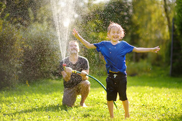 Funny little boy with his father playing with garden hose in sunny backyard. Preschooler child...