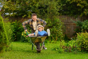 Fotobehang Happy little boy having fun in a wheelbarrow pushing by dad in domestic garden on warm sunny day. Active outdoors games for family with kids in the backyard in summer © Maria Sbytova