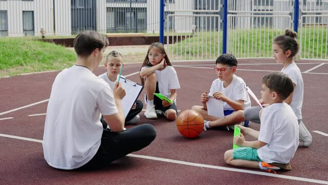 Male trainer explaining game strategy to kids on the basketball court. Diverse kids team and coach discussing game tactics. Education and sport concept. Back view.