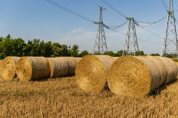 Round bales of straw are collected in a row near a high-voltage power line VL-750 for transportation. Metal supports in endless field. Blurred background. Selective focus. Nature concept for design.