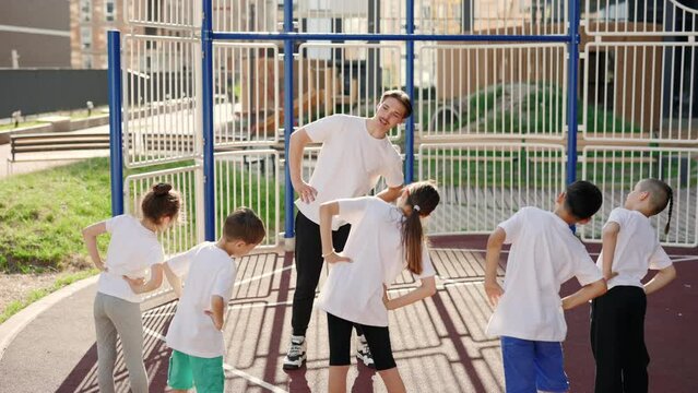 Male coach and primary school children do warm up before the workout outdoor. Group of elementary students exercising during class at school yard. Healthy sport concept.