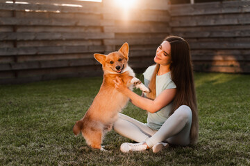 Domestic Corgi dog with girl owner. Young woman sitting on the grass and hug Welsh Corgi Pembroke. Lifestyle with domestic playful dog.