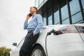 Handsome man in business suit surfing internet on modern smartphone while waiting electric car to...