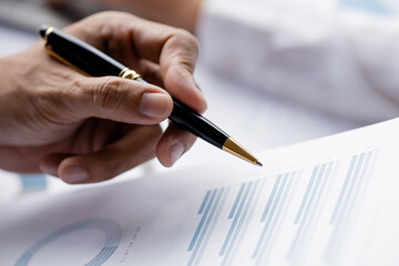 A close-up view of a businessman holding a pen pointing at a bar chart on a company financial document prepared by the Finance Department for a meeting with business partners. Financial concept.