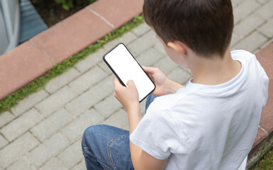 The boy is sitting on the street and holding a phone with a white empty screen in his hands and...