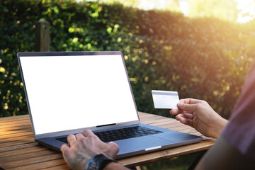 Young man using his credit card to pay online on his computer close-up view. Digital nomad working outside with white computer screen mockup