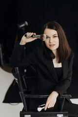 A girl in a black suit with a brush in her hands on a black background sits on a chair