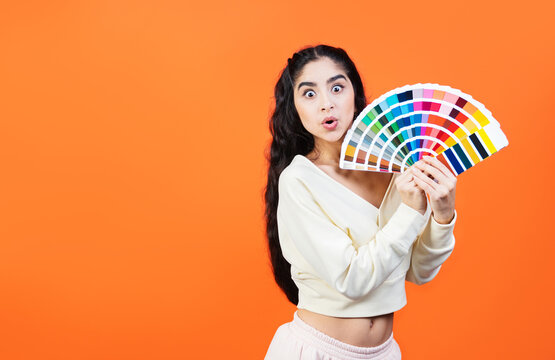 Very surprised woman with color palette fun. Paint and painting concept. Interior designer woman holding color palette guide on orange background with copy space.