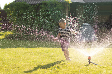 A little wet boy runs barefoot on the lawn next to the sprinkler. The concept of a happy, carefree childhood and holidays