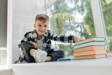 Fototapeta na wymiar A schoolboy sits on the windowsill with books to read