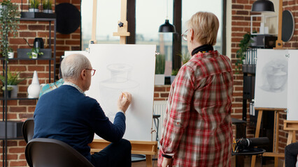 Artist supervising senior man drawing on canvas in art class, attending educational workshop to...