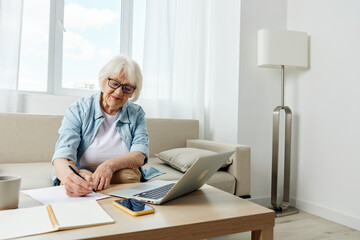 an elderly woman in stylish clothes is sitting on a sofa in a comfortable bright apartment and working on a laptop makes notes in a notebook. The concept of working from home