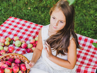 A little girl is sitting in the garden in the garden with a basket of ripe apples.