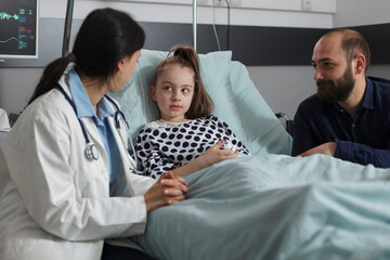 Pediatrician discussing with sick little daughter resting in patient bed while caring father sitting beside her. Pediatric healthcare facility medic expert consulting unwell girl health condition.