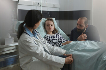 Pediatrician examining sick girl disease symptoms while resting in patient bed. Doctor expert discussing with girl about her health condition while caring father sitting beside ill daughter.