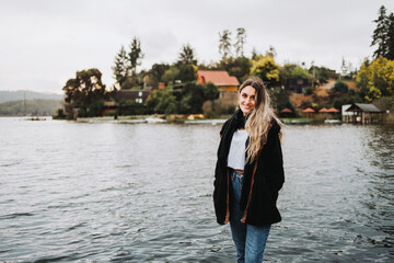 Young blonde warm woman standing and smiling in front of a beautiful lake on a cloudy afternoon. Copy space