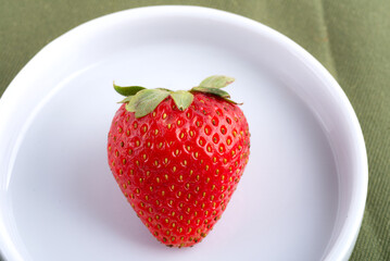 large red strawberries with green leaves in a white porcelain plate.