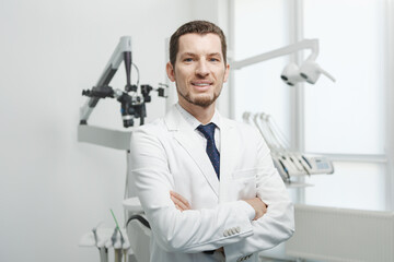 Naklejka na ściany i meble Happy male dentist in white lab coat posing with hands crossed and looking at camera