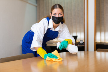 Fototapeta na wymiar Portrait of a young woman worker of a cleaning company in a protective mask, washing an office desk with a rag and detergent