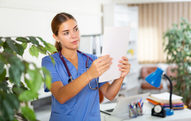 Young female doctor in surgical scrubs standing in hall of clinic. She's holding document in hands.