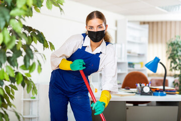 Young female worker of a cleaning company in a protective mask conducts a wet cleaning of the...