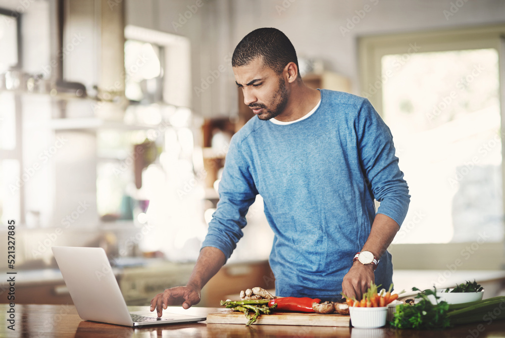 Poster young man using a laptop while cooking in a kitchen, checking the internet. male watching a tutorial