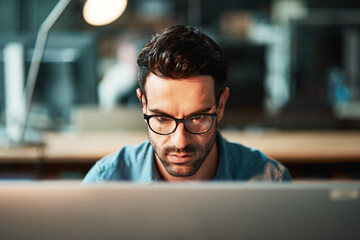 Serious business man working at computer late in the office at night to finish reports, articles or code. Focused and young male IT worker wearing glasses late in the evening for deadline