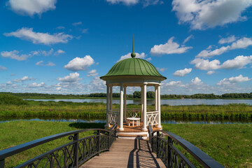 View of the gazebo with a bridge on the bank of the Volkhov River in the Novgorod Museum of Folk Wooden Architecture Vitoslavlitsa on a sunny summer day, Veliky Novgorod, Russia - obrazy, fototapety, plakaty