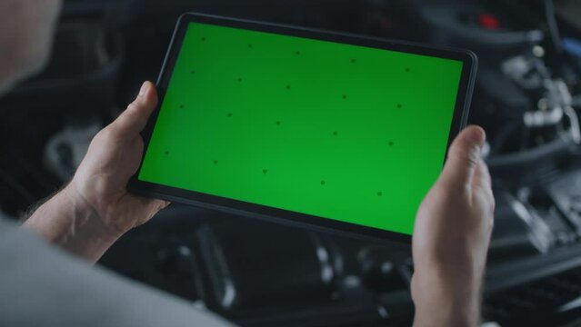 Unrecognizable male mechanic swiping on a tablet computer with chromakey green screen, and checking the results of car diagnostics while standing near the opened hood in a car service