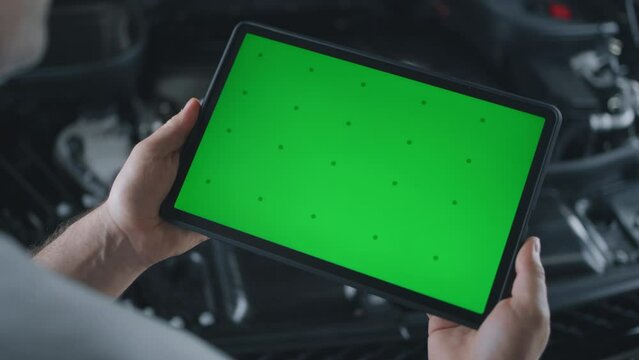 Unrecognizable mechanic holding a tablet with chromakey green screen in his hands, and standing near a car with an opened hood in a car service during a diagnostics