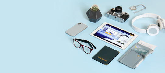 Open page of online booking service on screen of tablet computer and travel accessories on blue background with space for text
