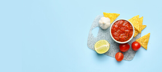 Bowl of tasty salsa sauce with ingredients and nachos on blue background with space for text