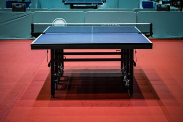 Blue table tennis or pingpong table is settle on a red, orange floor of the indoor court stadium for competitions tournament. - obrazy, fototapety, plakaty