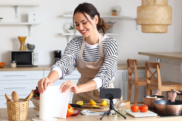 Young woman taking paper towel while following cooking video tutorial in kitchen - obrazy, fototapety, plakaty